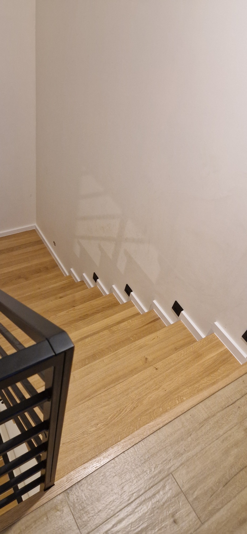 Widok z góry na drewniane schody z czarnymi akcentami, kontrastujące z jasną ścianą i podłogą, fragment czarnej barierki widoczny w lewym dolnym rogu.