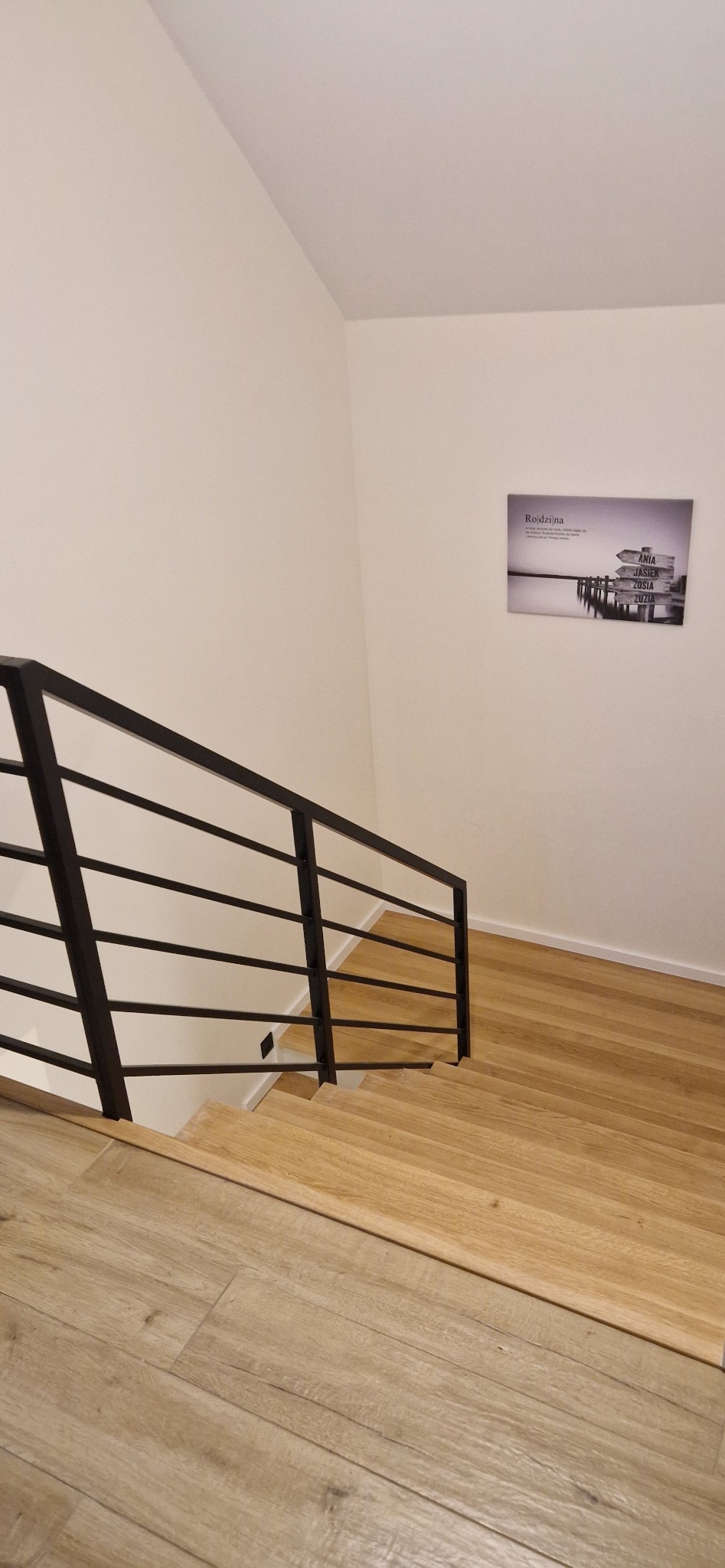Jasne, drewniane stopnie schodów z czarną, metalową balustradą, widok z góry, na ścianie wisi obrazek z motywem rodziny.