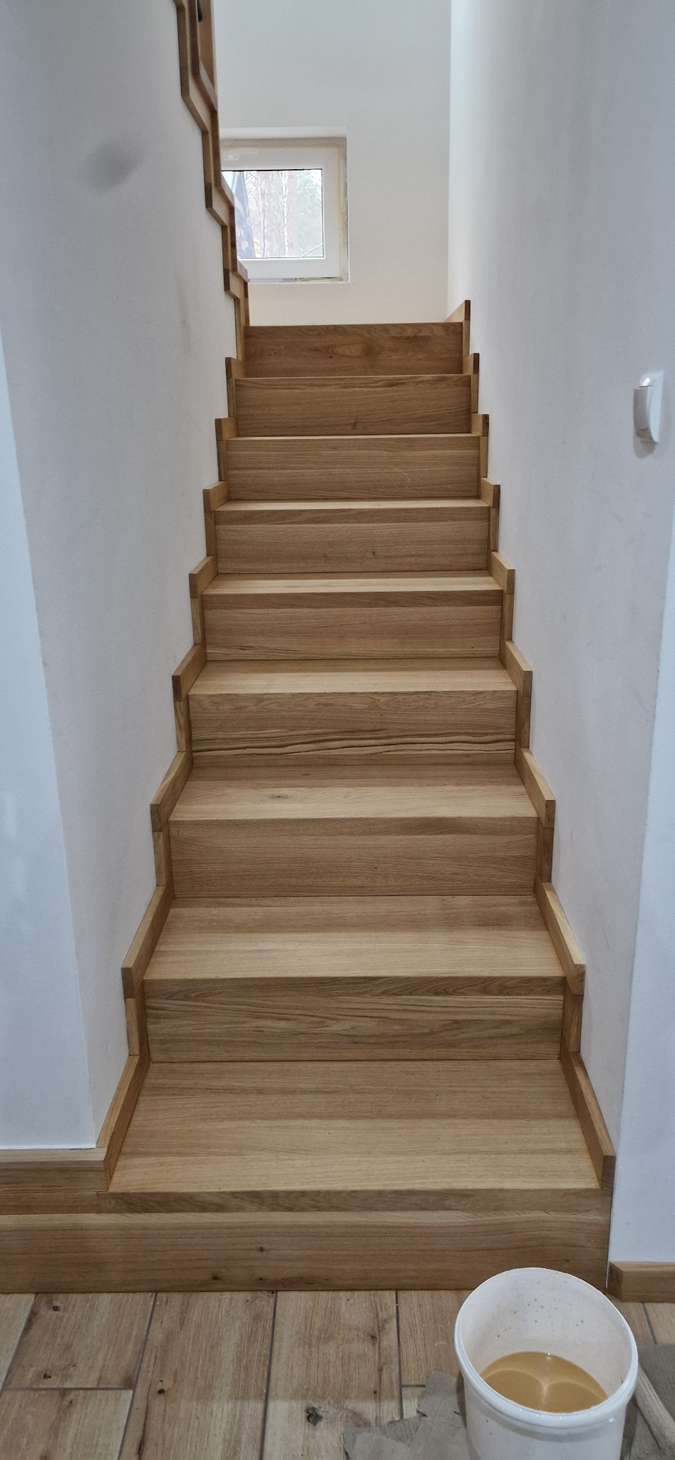 Jasne, dębowe schody z prostymi stopniami i podstopnicami, prowadzące w górę wzdłuż białej ściany, widoczne z dołu, z wiadrem farby na podłodze.
