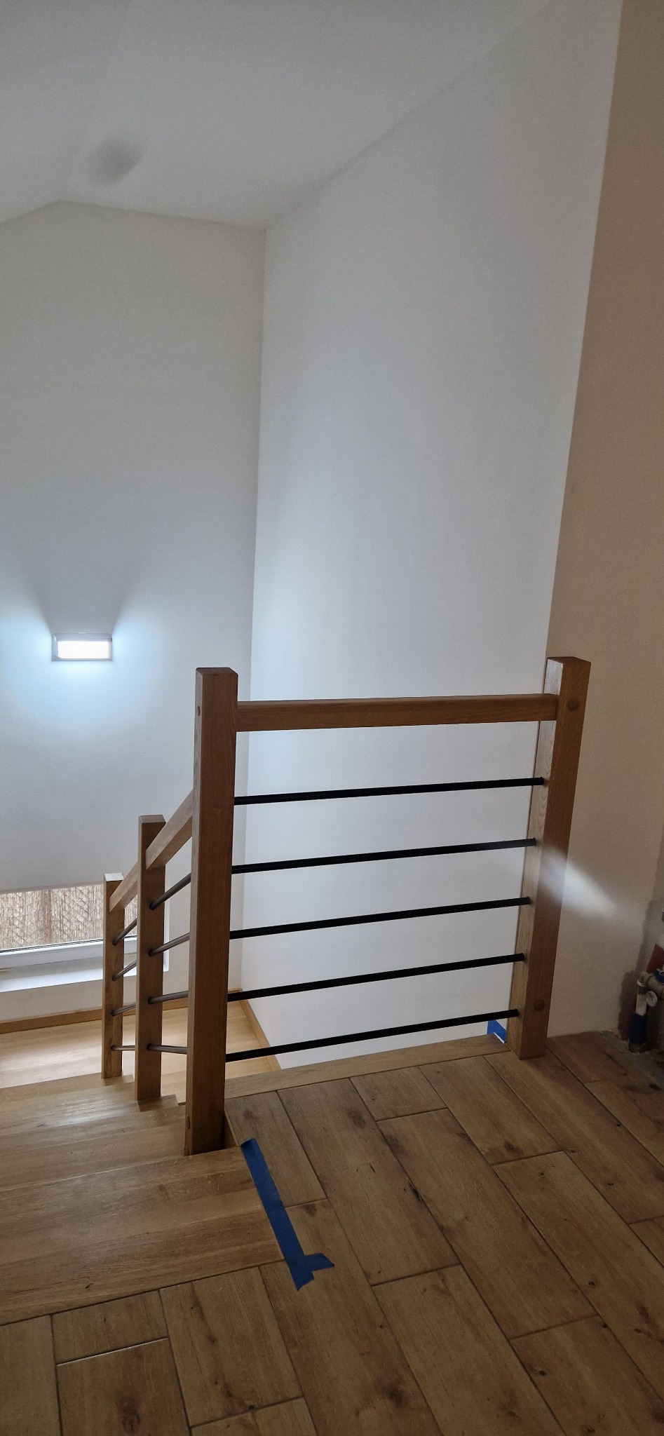 Fragment schodów z drewnianymi stopniami i balustradą z drewnianymi słupkami oraz czarnymi, poziomymi prętami, widok z góry na stopnie, zabezpieczone niebieską taśmą malarską.