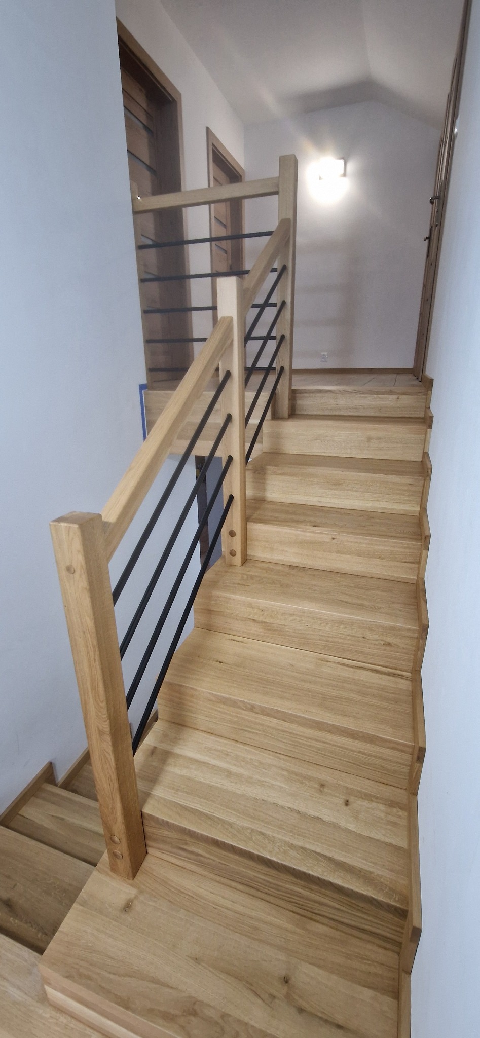 Jasne, drewniane schody z minimalistyczną balustradą z czarnych, poziomych prętów, prowadzące na górę, widok z dołu.