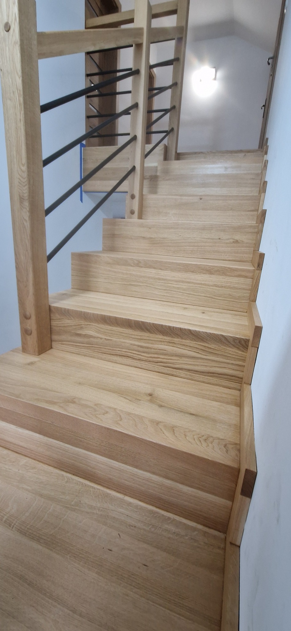 Jasne, dębowe schody z czarnymi metalowymi balustradami, widok z dołu, ukazujący strukturę drewna i minimalistyczny design.