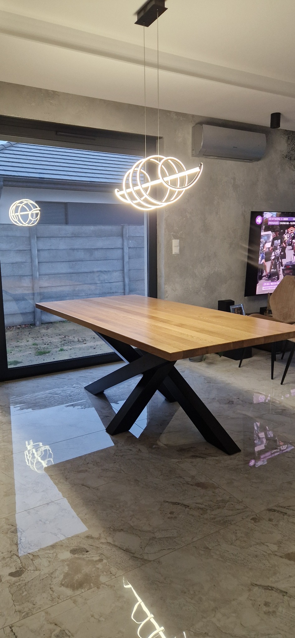 Stół drewniany z blatem z jasnego drewna i czarnymi nogami w kształcie litery X, oświetlony nowoczesną lampą wiszącą z jasnymi okręgami, na tle betonowej ściany i okna z widokiem na betonowy płot.