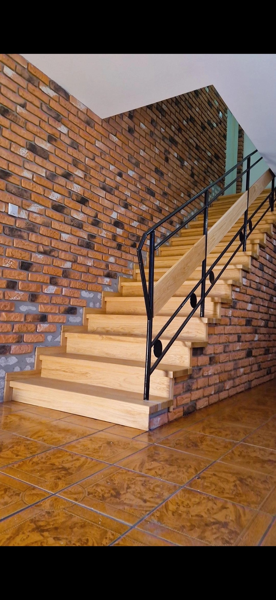 Jasne, drewniane stopnie schodów z czarną, metalową balustradą, przylegające do ściany wyłożonej cegłą klinkierową, na brązowej, błyszczącej podłodze.