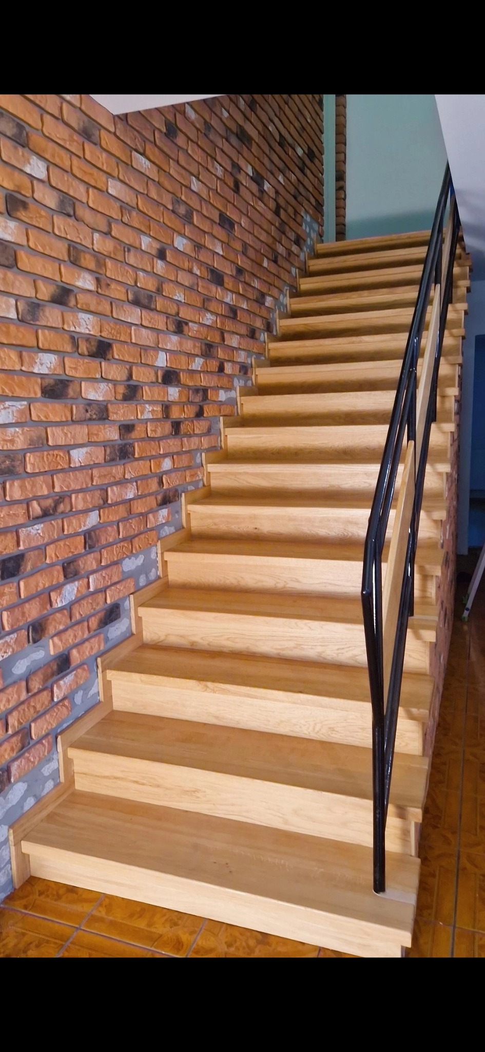 Jasne, drewniane schody z czarną, metalową balustradą w domu z ceglaną ścianą.