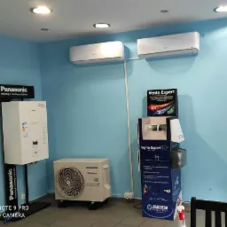 Ekspozycja w salonie: Dwa klimatyzatory Panasonic zamontowane na ścianie, obok jednostka zewnętrzna i model grzewczy na stojaku, uzupełnione o stand reklamowy Vento Expert i logo Blauberga.