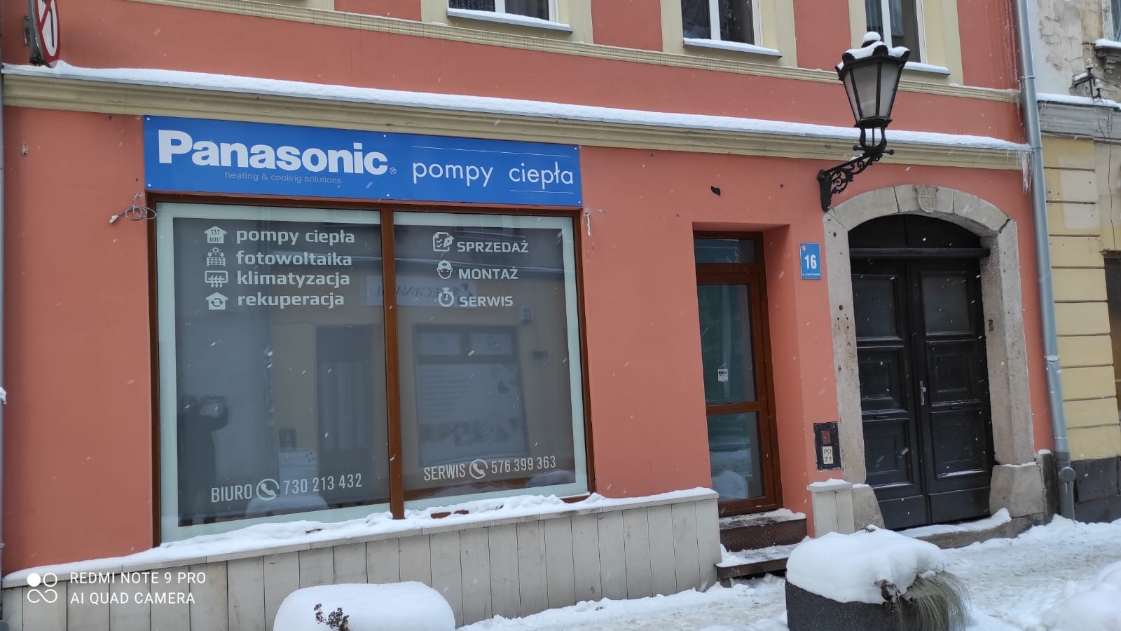 Fasada budynku w zimowej scenerii z szyldem Panasonic i informacjami o sprzedaży, montażu i serwisie pomp ciepła, fotowoltaiki, klimatyzacji i rekuperacji. Widoczny numer budynku 16 i zabytkowa...