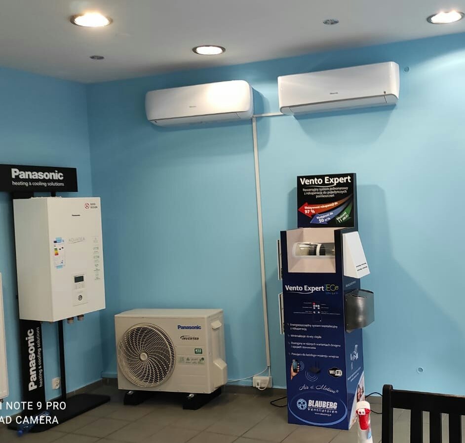 Ekspozycja w salonie: Dwa klimatyzatory Panasonic zamontowane na ścianie, obok jednostka zewnętrzna i model grzewczy na stojaku, uzupełnione o stand reklamowy Vento Expert i logo Blauberga.
