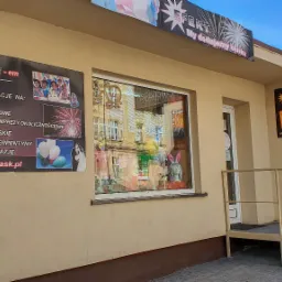 Zewnętrzny widok na budynek firmy oferującej artykuły i dekoracje na różne okazje, w tym fajerwerki i balony, z widocznymi szyldami reklamowymi i wystawą w oknie.