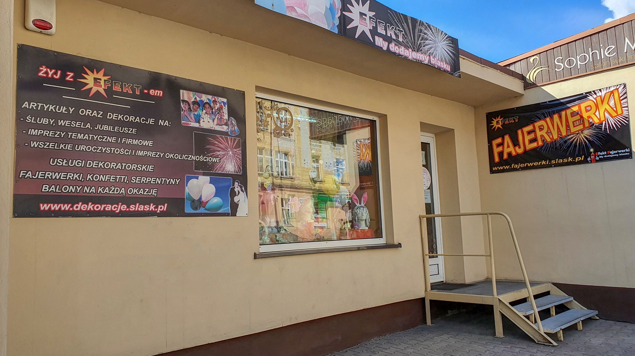Zewnętrzny widok na budynek firmy oferującej artykuły i dekoracje na różne okazje, w tym fajerwerki i balony, z widocznymi szyldami reklamowymi i wystawą w oknie.