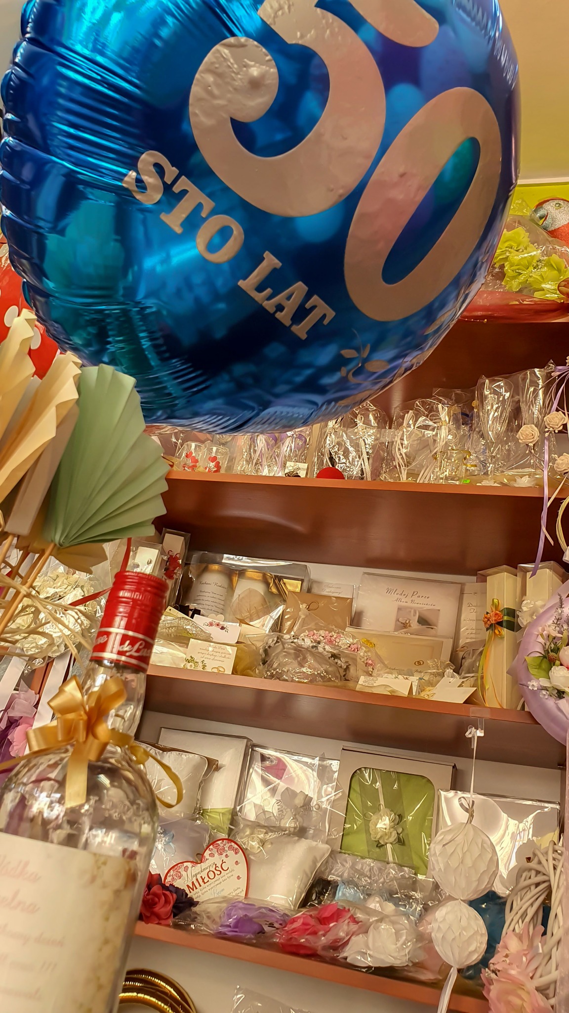 Sklep z akcesoriami imprezowymi: niebieski balon foliowy z napisem 'STO LAT 50', ozdobna butelka z kokardą, wachlarze papierowe i półki z zapakowanymi prezentami, kartkami okolicznościowymi...