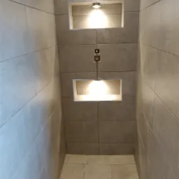 Wnętrze kabiny prysznicowej wyłożonej szarymi, podłużnymi płytkami ceramicznymi, z dwoma wnękami oświetlonymi punktowymi lampami, z widocznym systemem mocowania oświetlenia.