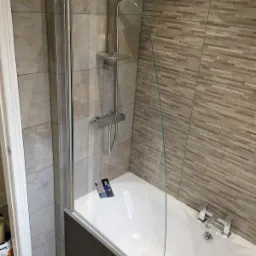 Wykończona łazienka w Ciechanowie, prezentująca wannę z zamontowanym szklanym parawanem nawannowym oraz nową armaturą i płytkami ściennymi w odcieniach beżu i brązu.