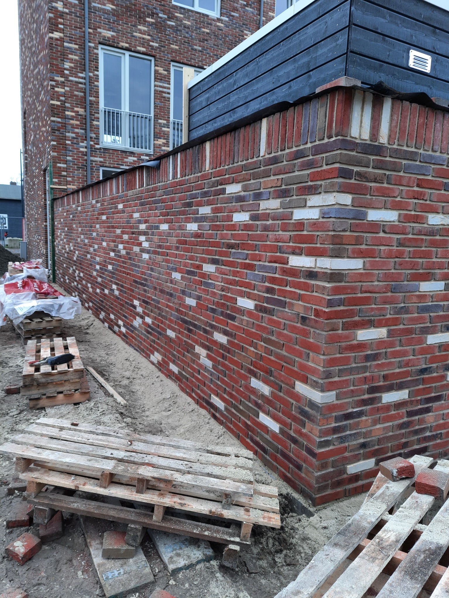 Wznoszenie muru z cegły klinkierowej w odcieniach czerwieni i bieli, widoczne palety i zapasy materiału budowlanego na placu budowy.