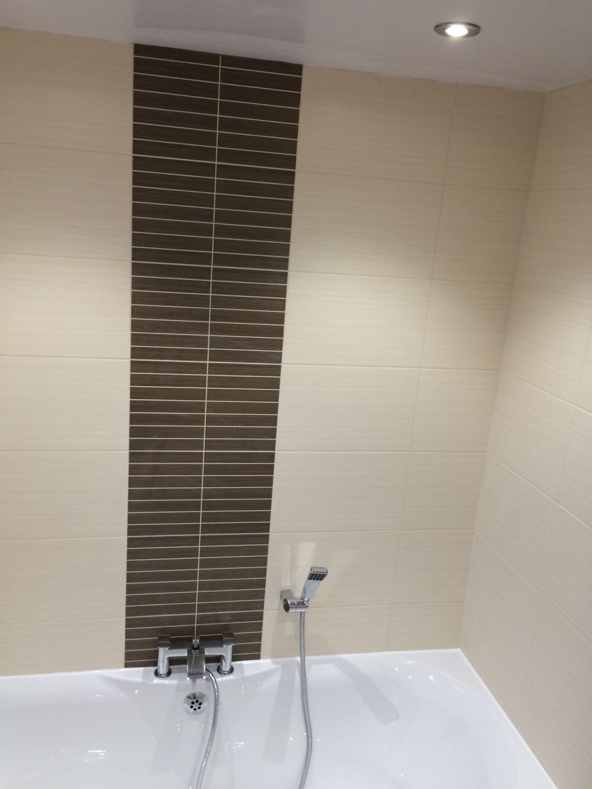 Wykończona łazienka z wanną i baterią, ściany wyłożone beżowymi płytkami z pionowym pasem płytek w kolorze ciemnego brązu, oświetlenie halogenowe w suficie.