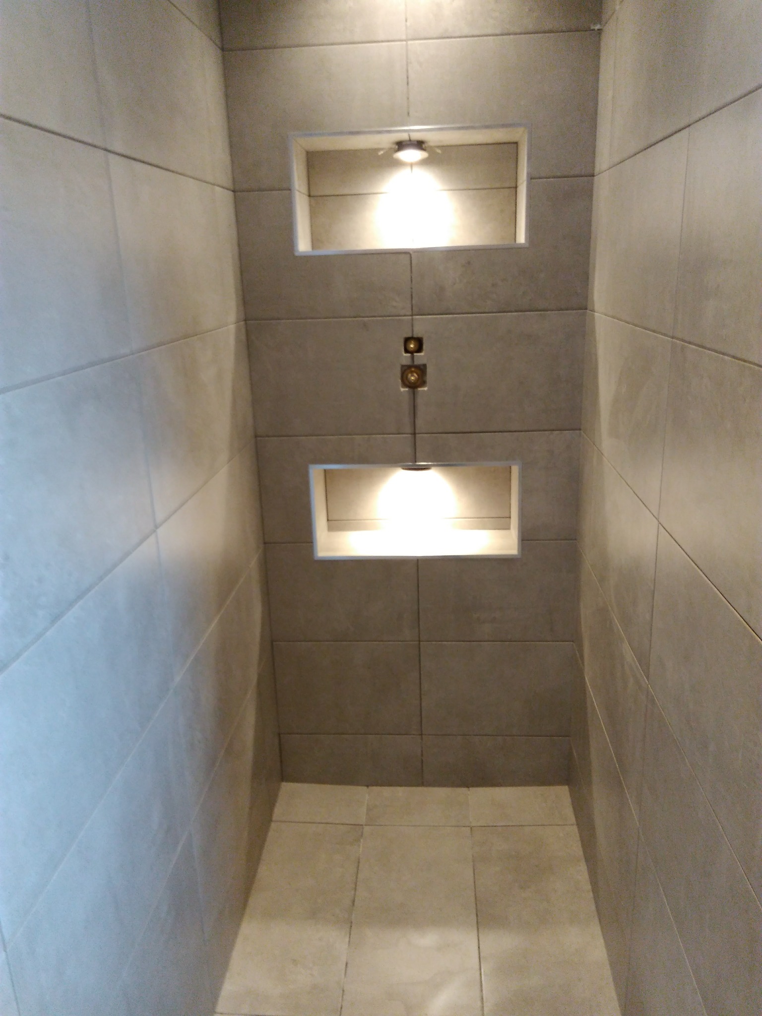 Wnętrze kabiny prysznicowej wyłożonej szarymi, podłużnymi płytkami ceramicznymi, z dwoma wnękami oświetlonymi punktowymi lampami, z widocznym systemem mocowania oświetlenia.