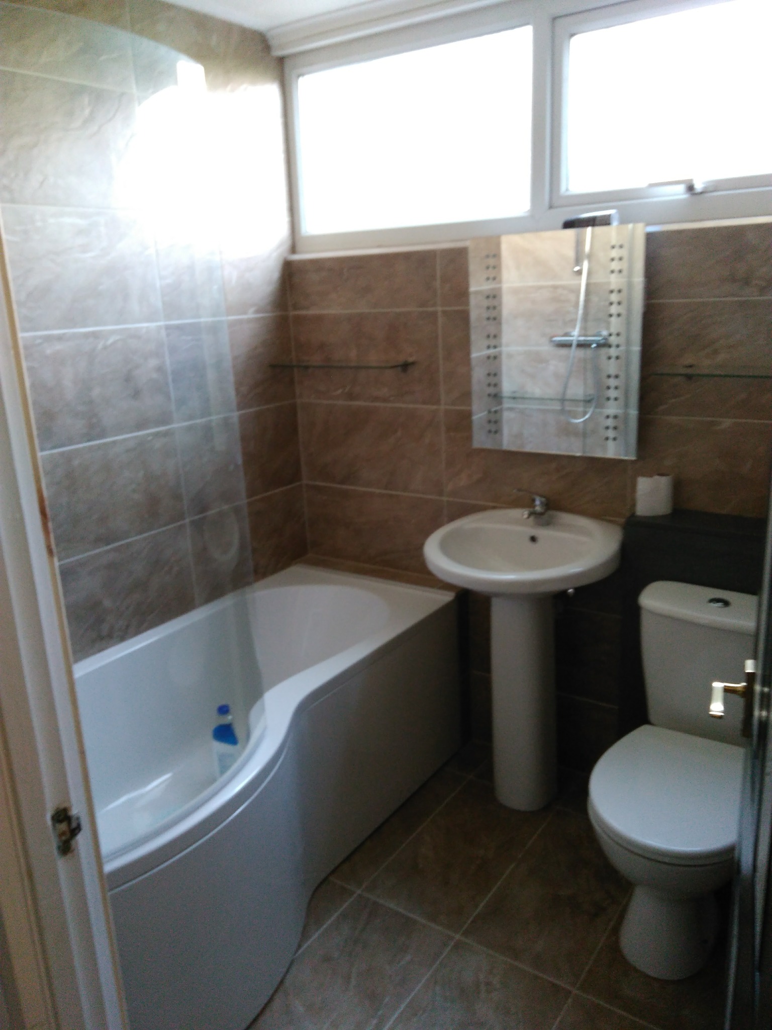 Wykończona łazienka z brązowymi płytkami, białą wanną asymetryczną, umywalką na postumencie i toaletą, oświetlona przez okno.