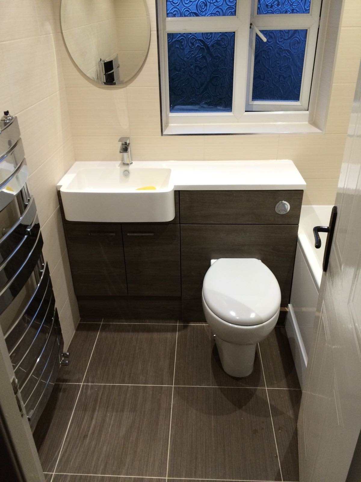 Nowoczesna łazienka z umywalką w zabudowie z szafką w kolorze ciemnego drewna, toaletą, grzejnikiem drabinkowym i oknem z ozdobną szybą.