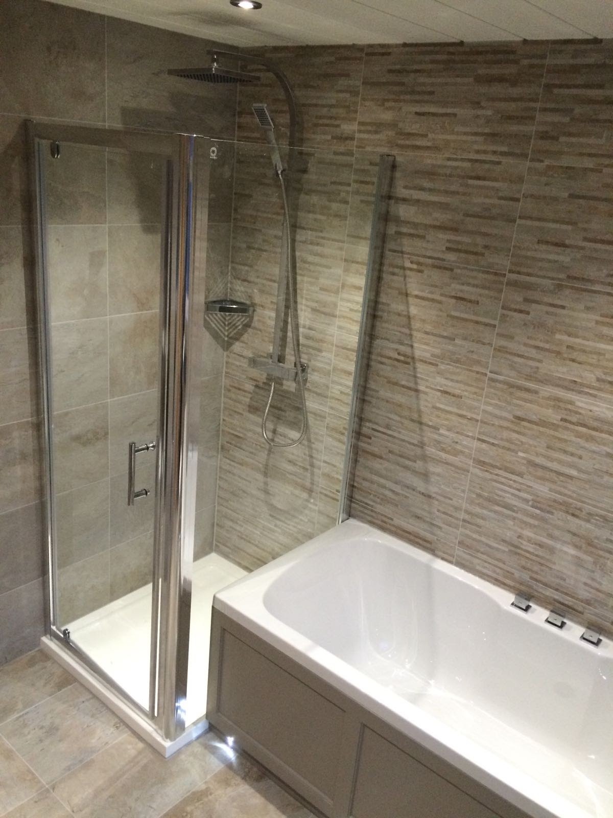 Nowoczesna łazienka z kabiną prysznicową ze srebrnymi ramami, deszczownicą i wanną obudowaną panelami w odcieniach szarości, wykończona płytkami w beżowo-brązowe paski.