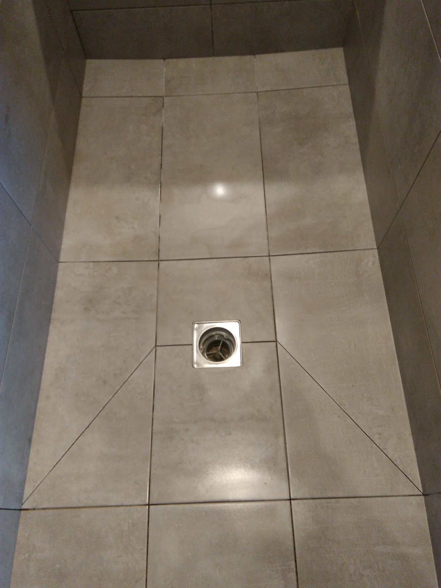 Wykończony brodzik z jasnoszarych, kwadratowych płytek ceramicznych, z centralnie umieszczonym odpływem liniowym w metalowej ramce i spadem w jego kierunku.