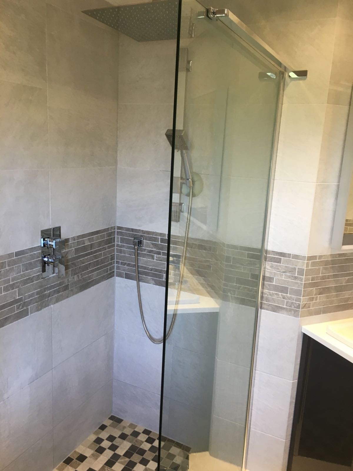 Nowoczesna kabina prysznicowa z deszczownicą, wykończona płytkami w odcieniach szarości z mozaiką na podłodze, widoczny chromowany panel sterujący i szklane drzwi przesuwne.