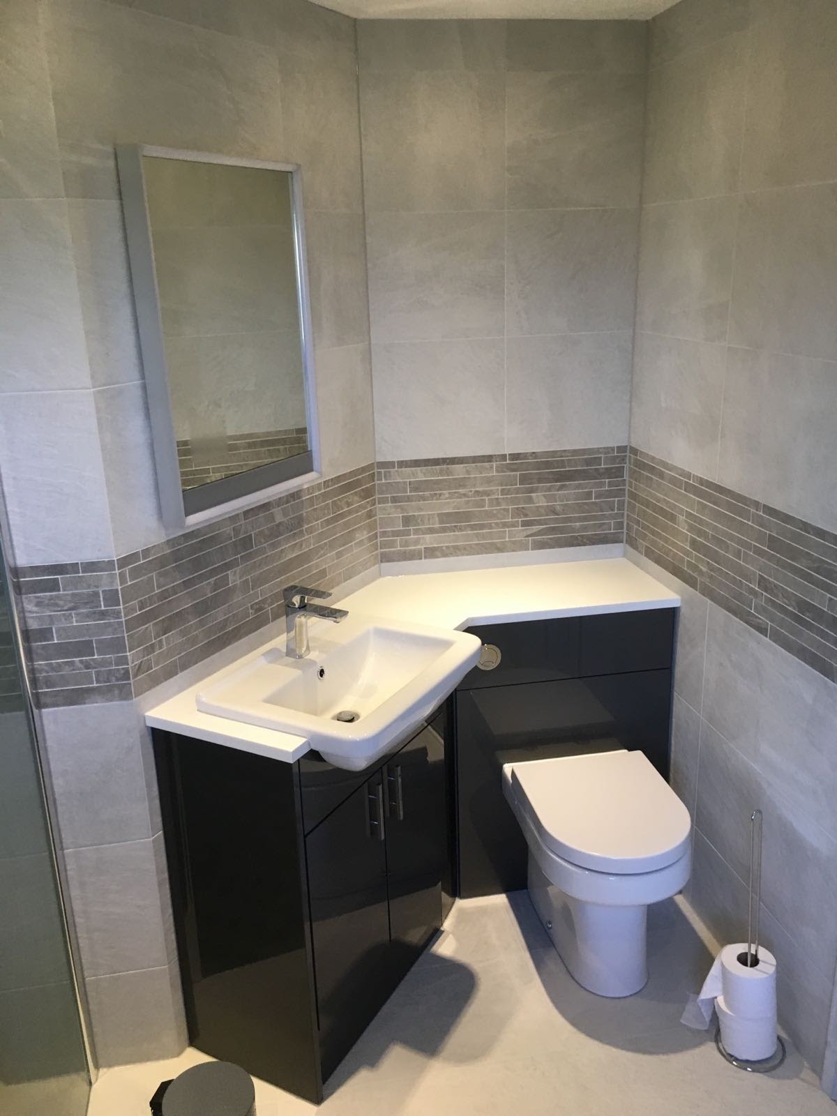 Ukończona łazienka z szarymi płytkami, umywalką w szafce z ciemnego lakierowanego drewna, toaletą i dekoracyjnym pasem z mozaiki.