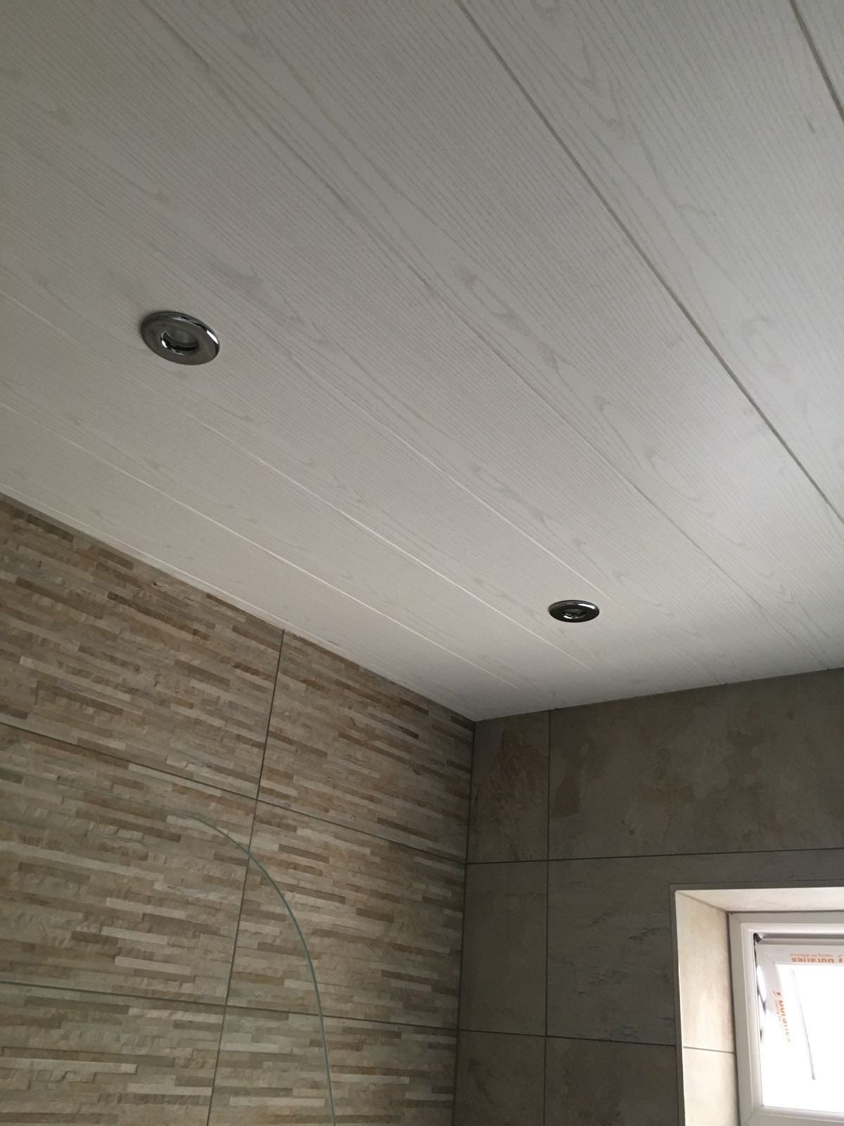 Wykończona łazienka z drewnopodobnym panelem sufitowym, w którym zamontowano dwa okrągłe halogeny, oraz ścianami wyłożonymi różnymi odcieniami płytek imitujących kamień.