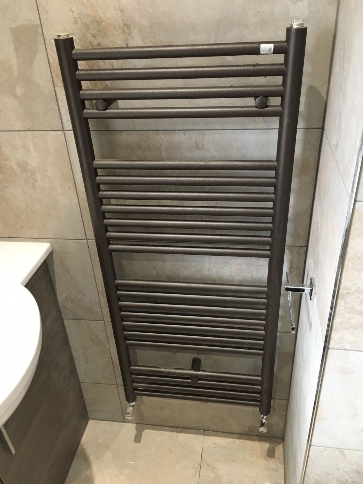 Brązowy grzejnik drabinkowy zamontowany na beżowych, kwadratowych płytkach w łazience, widoczny fragment umywalki po lewej stronie.