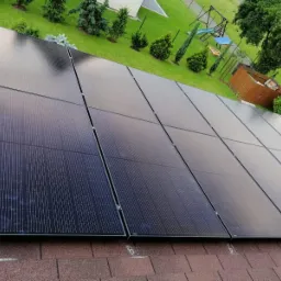 Czarny panel fotowoltaiczny na brązowym dachu z widokiem na zielony trawnik i drzewa.