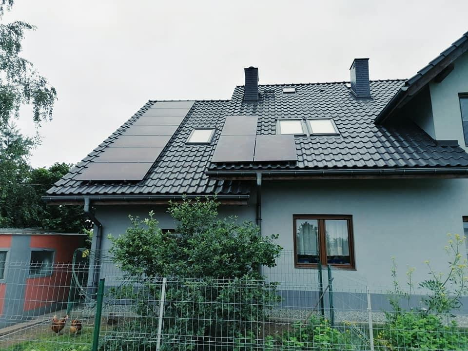 Dom jednorodzinny z panelami słonecznymi na dachu pokrytym ciemną dachówką, widoczne okna dachowe i komin, ogrodzenie z zielonej siatki, w tle kury.