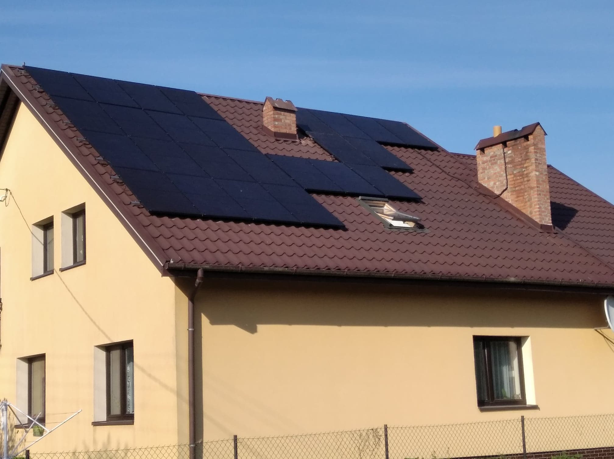 Dom jednorodzinny z brązowym dachem pokryty panelami słonecznymi, widoczne dwa kominy z cegły i okna z ciemnymi ramami.