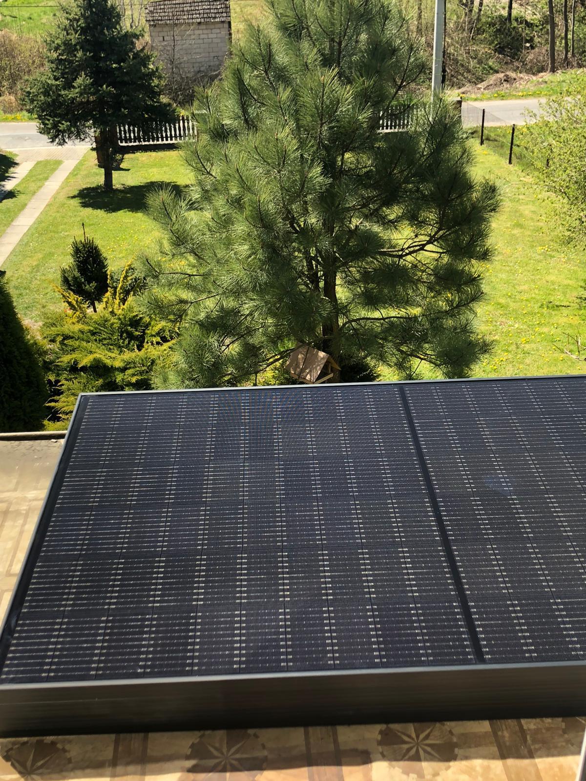 Panel słoneczny na dachu z widokiem na zielony ogród z drzewami iglastymi i budynkiem w tle.