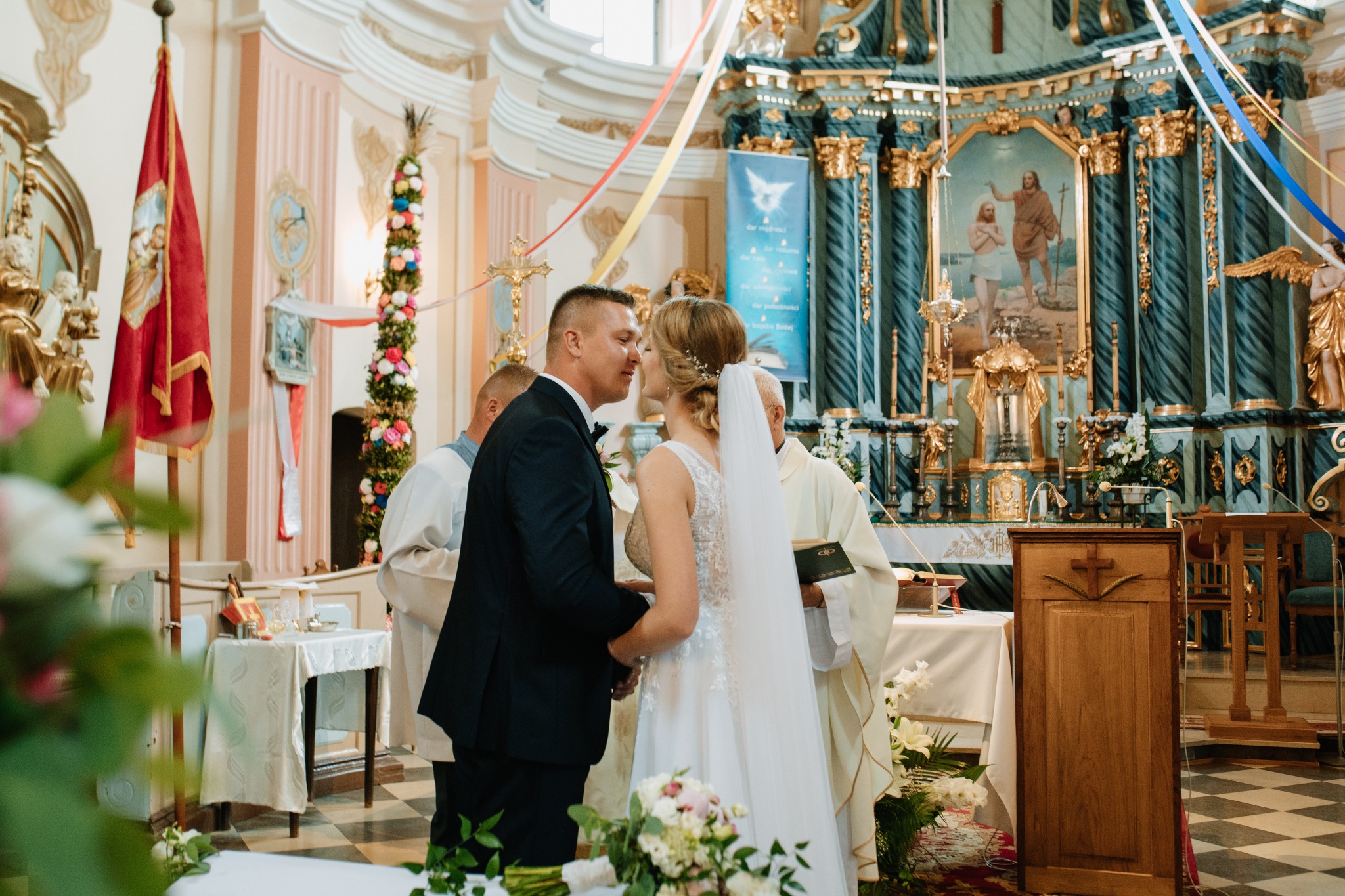 Pocałunek Pary Młodej w kościele podczas ceremonii ślubnej, w tle bogato zdobiony ołtarz i dekoracje kwiatowe.