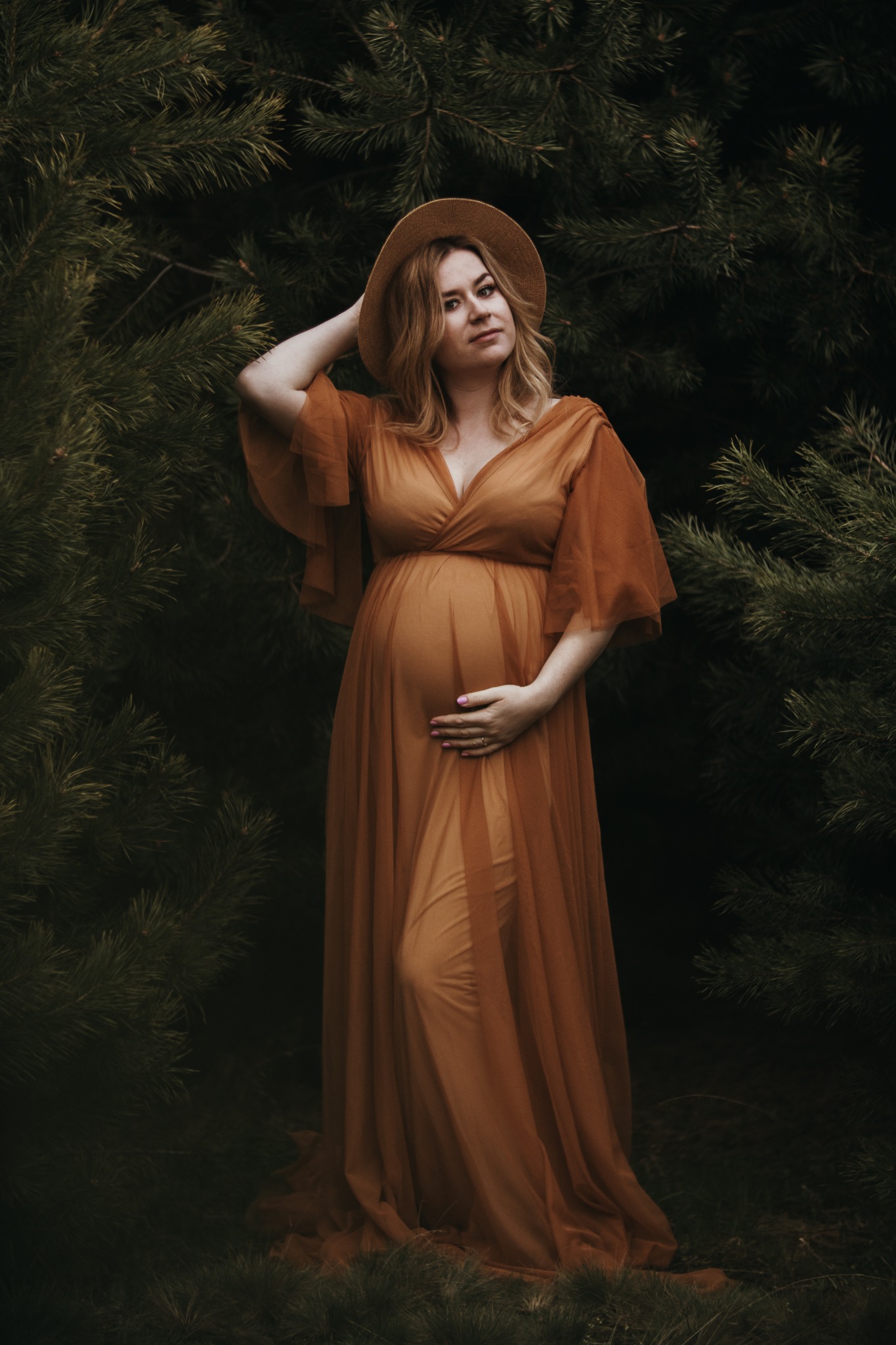 Kobieta w pomarańczowej, zwiewnej sukni i kapeluszu pozuje w otoczeniu ciemnych drzew iglastych, trzymając dłoń na brzuchu.