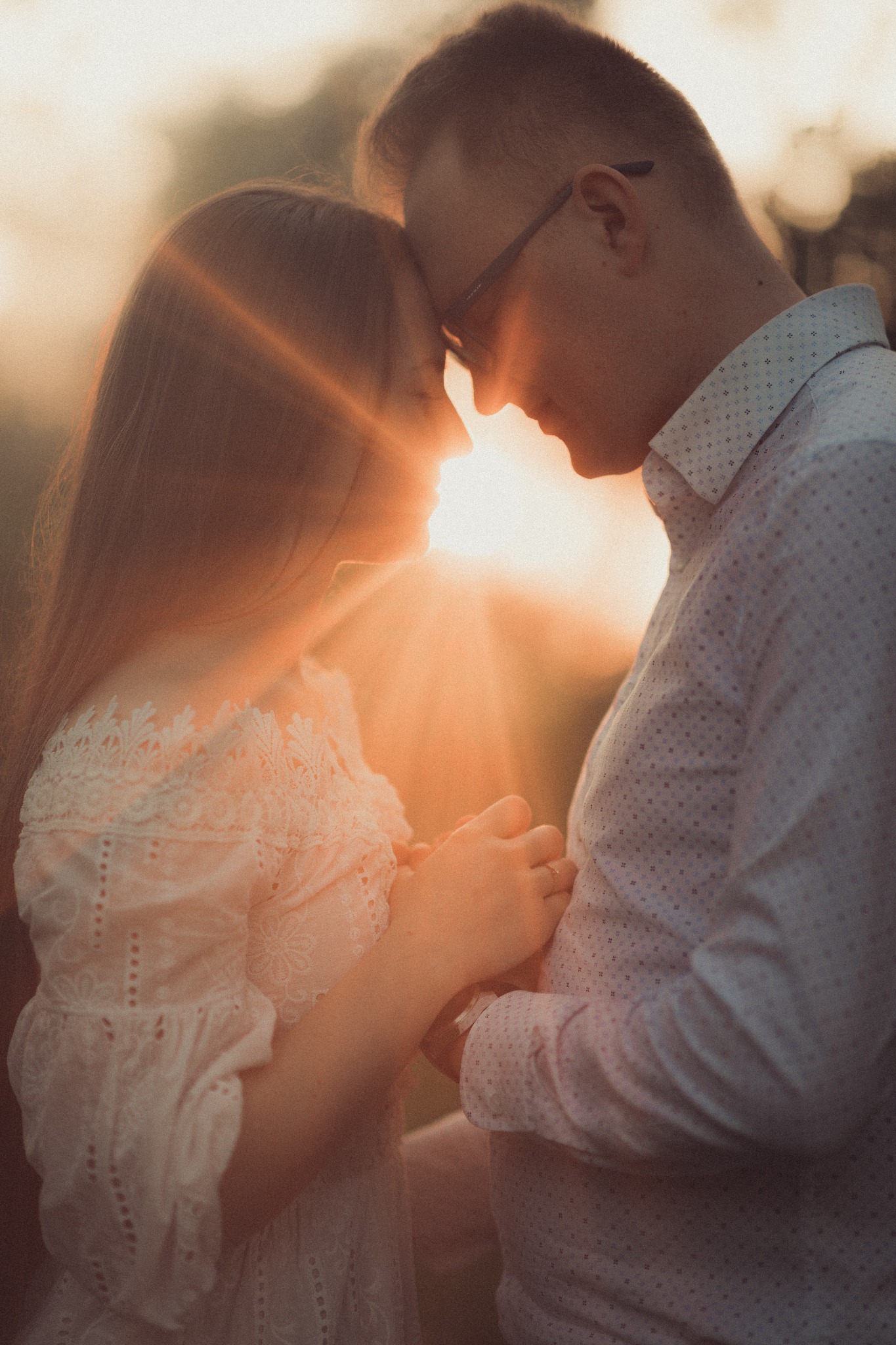 Zakochana para w kontraście ze słońcem, ona w koronkowej sukience, on w koszuli w drobny wzór, trzymają się za ręce, złączeni czołami.