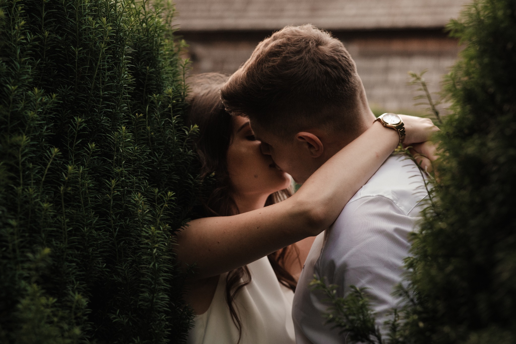 Pocałunek młodej pary uchwycony zza zielonych krzewów, mężczyzna w białej koszuli z zegarkiem na ręce obejmuje kobietę.