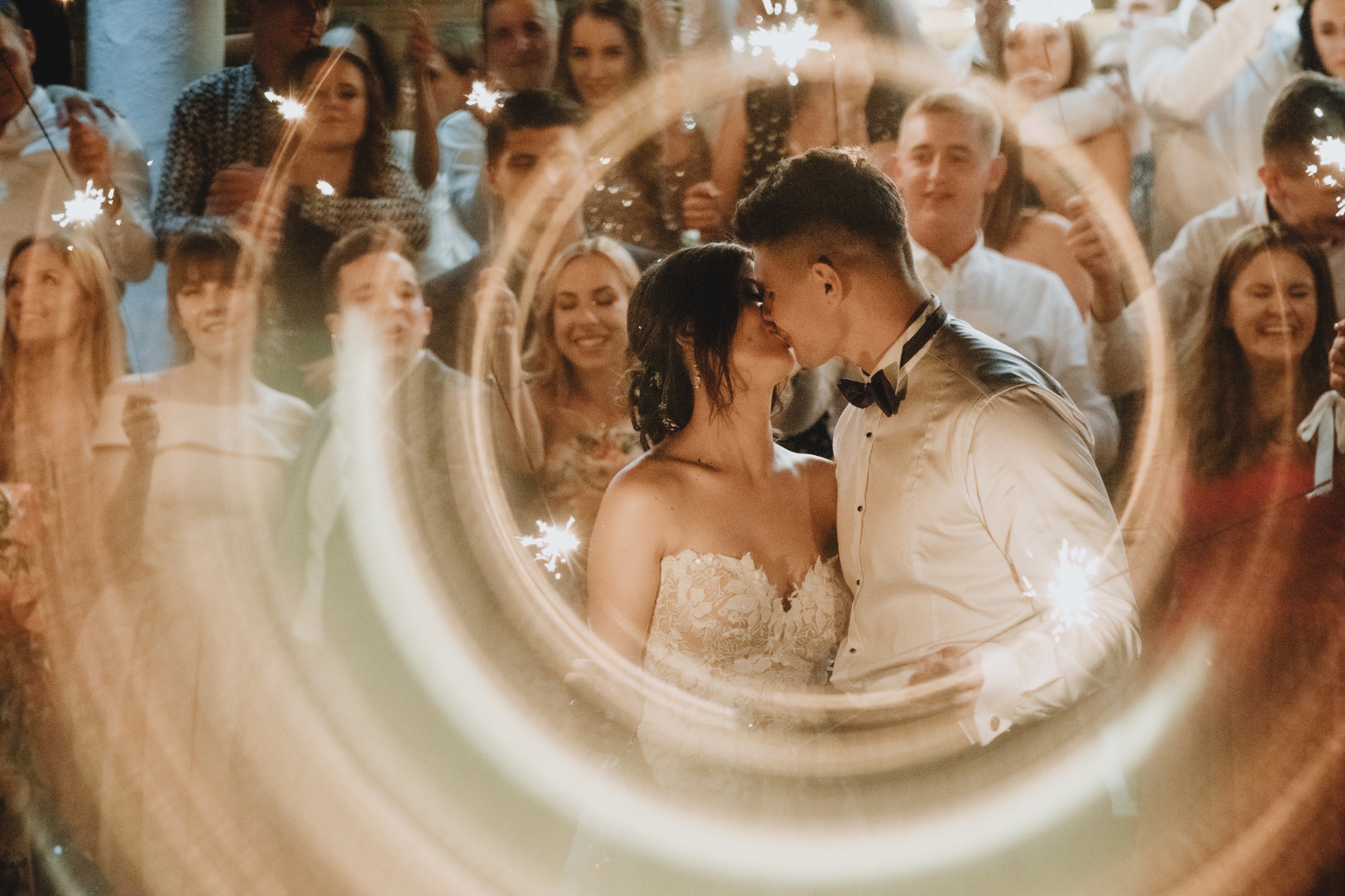 Pocałunek pary młodej otoczonej gośćmi z zimnymi ogniami, rozmyte światło tworzy efekt tunelu, panna młoda w koronkowej sukni bez ramiączek, pan młody w białej koszuli z muszką.