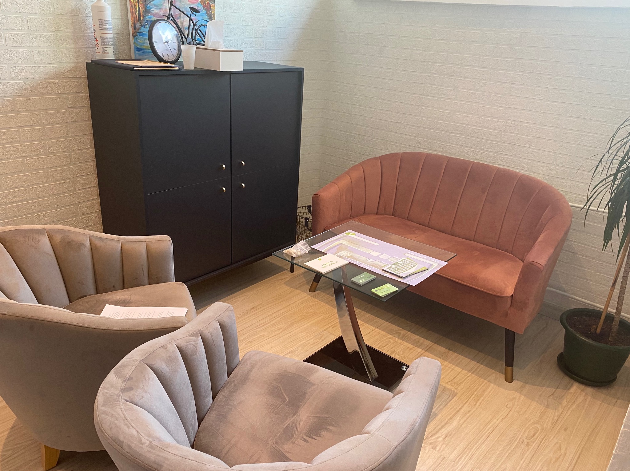 Jasne wnętrze gabinetu z dwoma beżowymi fotelami, brzoskwiniową sofą, szklanym stolikiem ze schematami i czarną szafką z dekoracjami na górze, tworzące przytulną przestrzeń do rozmów.