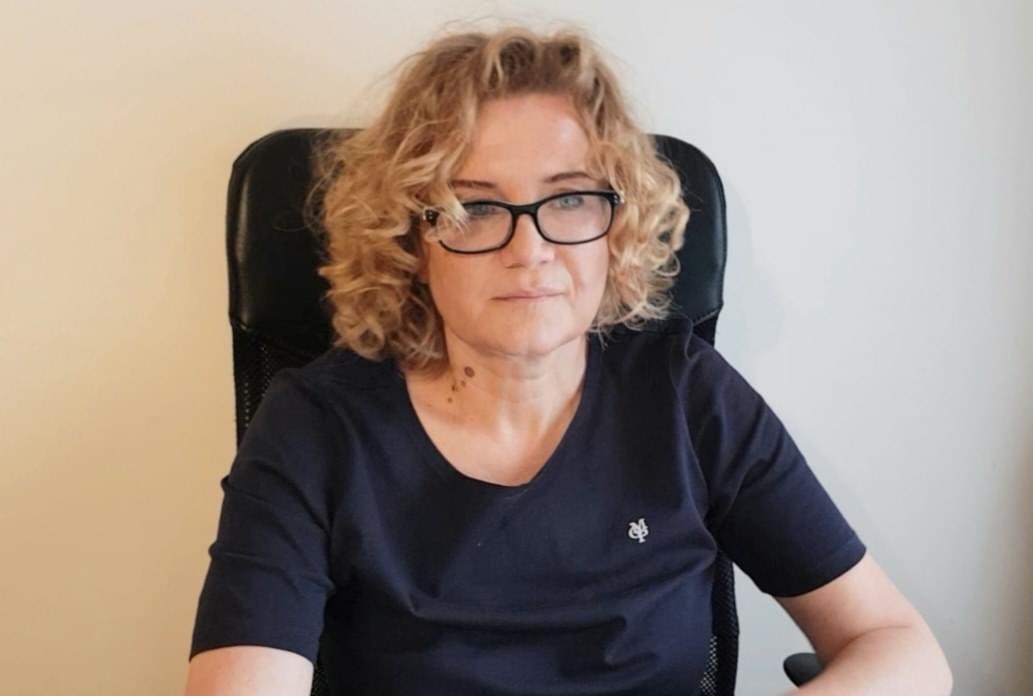 Portret kobiety z kręconymi blond włosami i okularach, siedzącej na czarnym krześle na tle jasnej ściany.