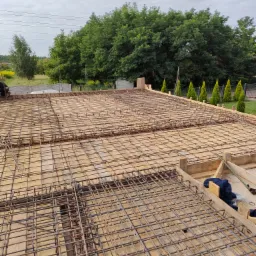 Siatka zbrojeniowa na szalunku z desek, przygotowana pod wylewkę betonową; w tle drzewa i błękitne niebo.