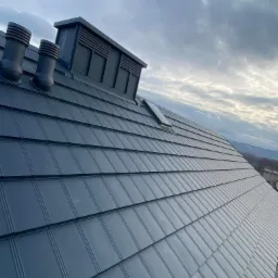 Szczegółowe ujęcie nowo położonego, grafitowego dachu z blachy na rąbek stojący, z widocznymi kominkami wentylacyjnymi i oknem dachowym, na tle pochmurnego nieba.