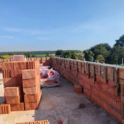Wznoszenie ścian zewnętrznych z czerwonej cegły ceramicznej szczelinowej, wzmocnionej drewnianym szalunkiem z metalowymi ściągami, widok na okoliczny krajobraz pod błękitnym niebem.