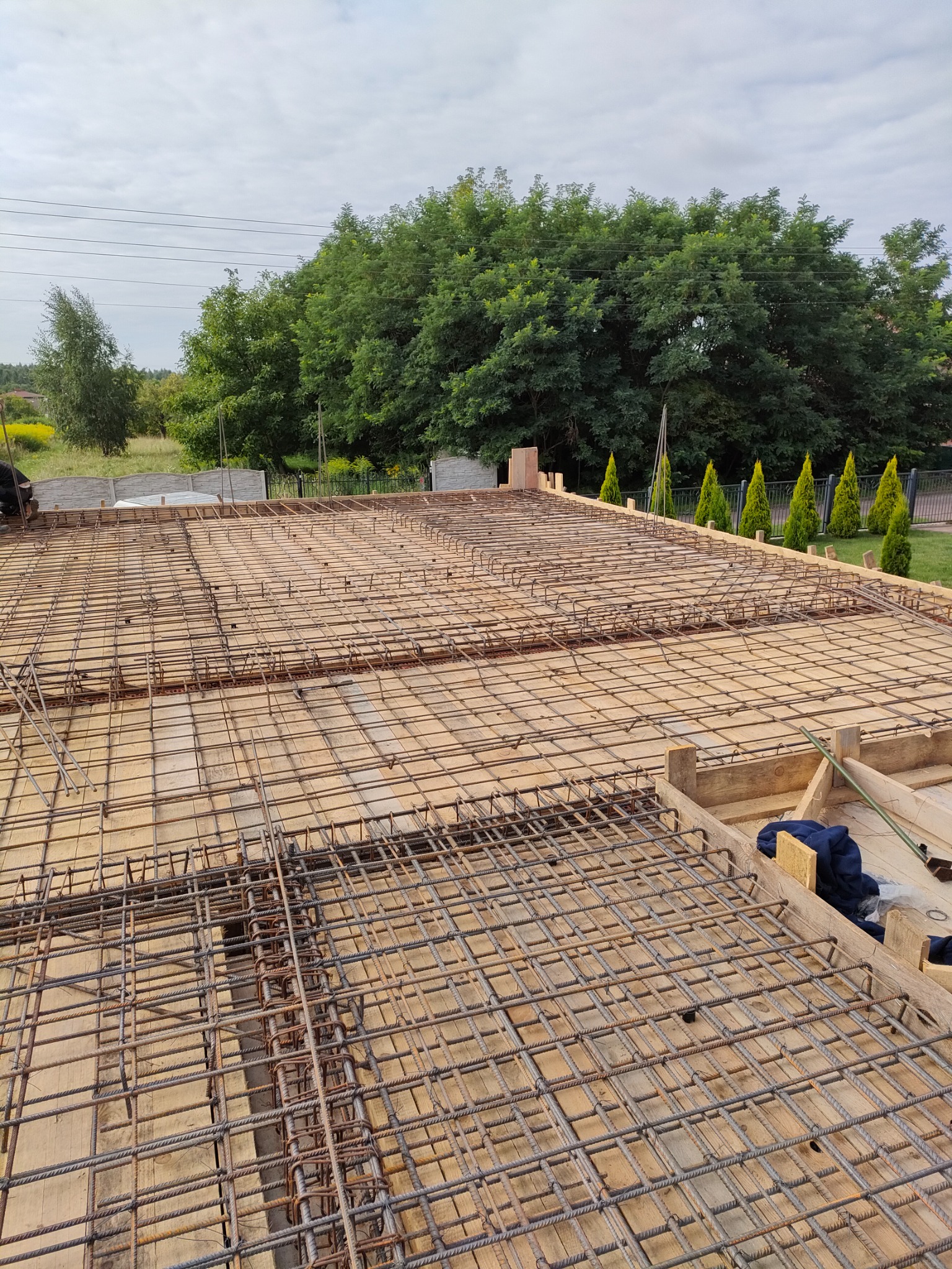 Siatka zbrojeniowa na szalunku z desek, przygotowana pod wylewkę betonową; w tle drzewa i błękitne niebo.