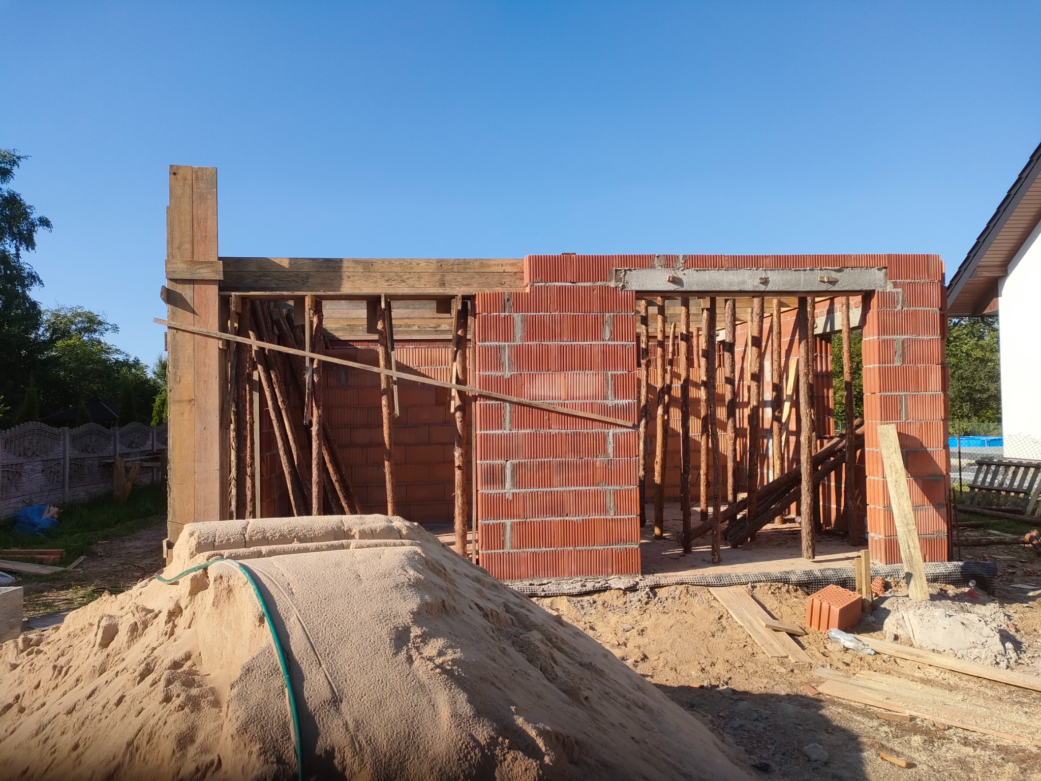 Wznoszenie ścian z czerwonej cegły, widoczne drewniane szalunki i sterta piasku z zielonym wężem na placu budowy w słoneczny dzień.