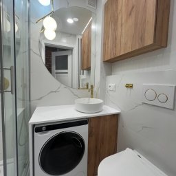 ŁAZIENKA W2 TYGODNIE Andrzej Kukla - Nowoczesna łazienka z marmurowymi płytkami, okrągłym lustrem, drewnianymi szafkami i pralką w zabudowie pod umywalką. Złote akcenty i oświetlenie LED.