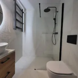 Minimalistyczna łazienka z kabiną typu walk-in, czarną armaturą i drewnianą szafką pod umywalką. Ściany i podłoga wyłożone płytkami imitującymi marmur.