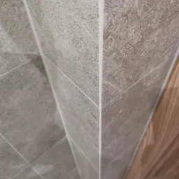 Szare płytki gresowe imitujące kamień na ścianie, z widoczną fugą i fragmentem drewnianej ościeżnicy drzwiowej w rogu.