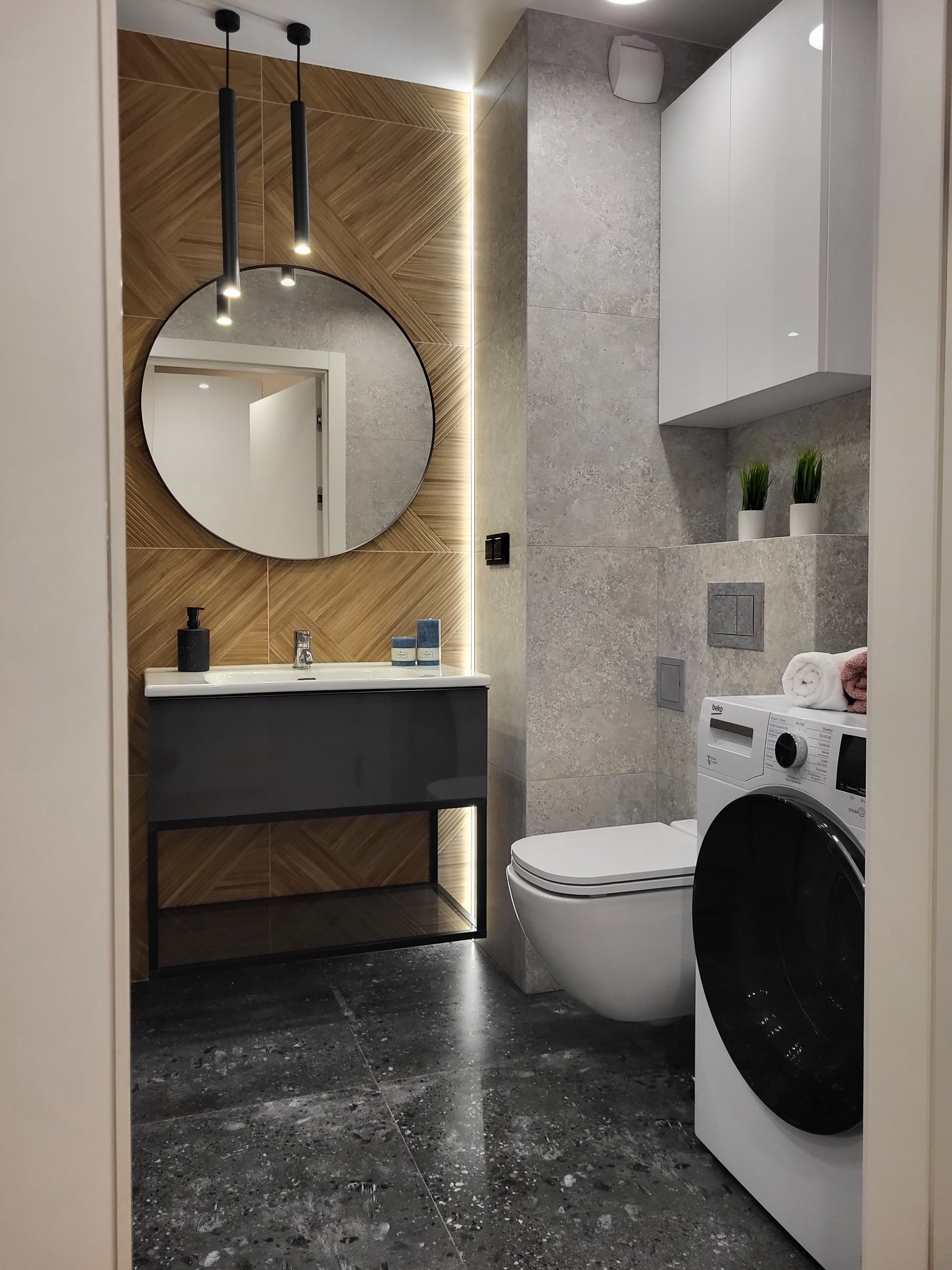 Nowoczesna łazienka z drewnianą ścianą za okrągłym lustrem, szarą umywalką, toaletą i pralką. Minimalistyczne lampy wiszące i szare płytki podłogowe.