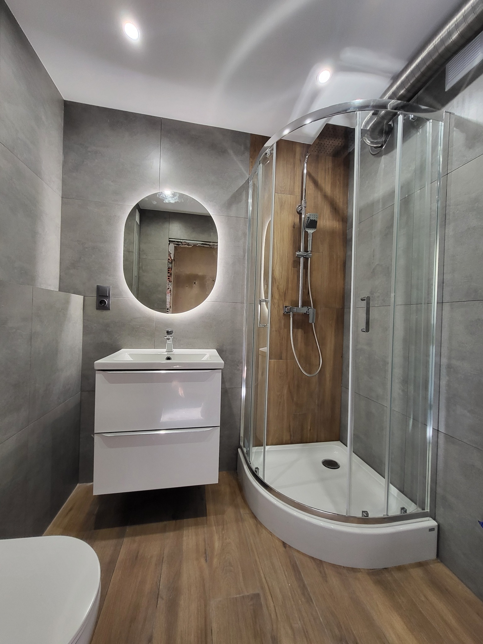 Nowoczesna łazienka: szare płytki, drewnopodobna podłoga, okrągłe lustro z podświetleniem i przeszklona kabina prysznicowa. Minimalistyczny styl.