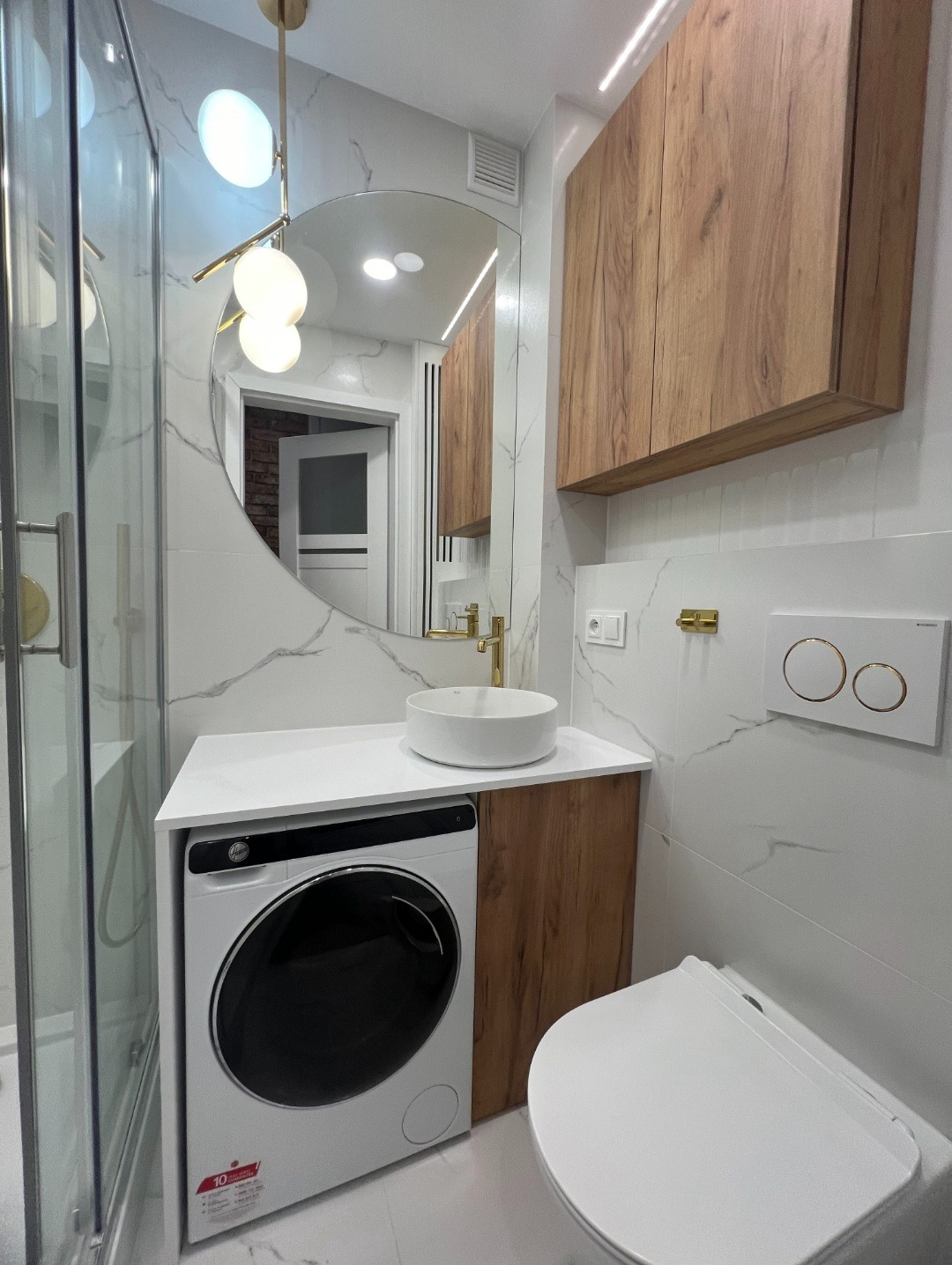 Nowoczesna łazienka z marmurowymi płytkami, okrągłym lustrem, drewnianymi szafkami i pralką w zabudowie pod umywalką. Złote akcenty i oświetlenie LED.